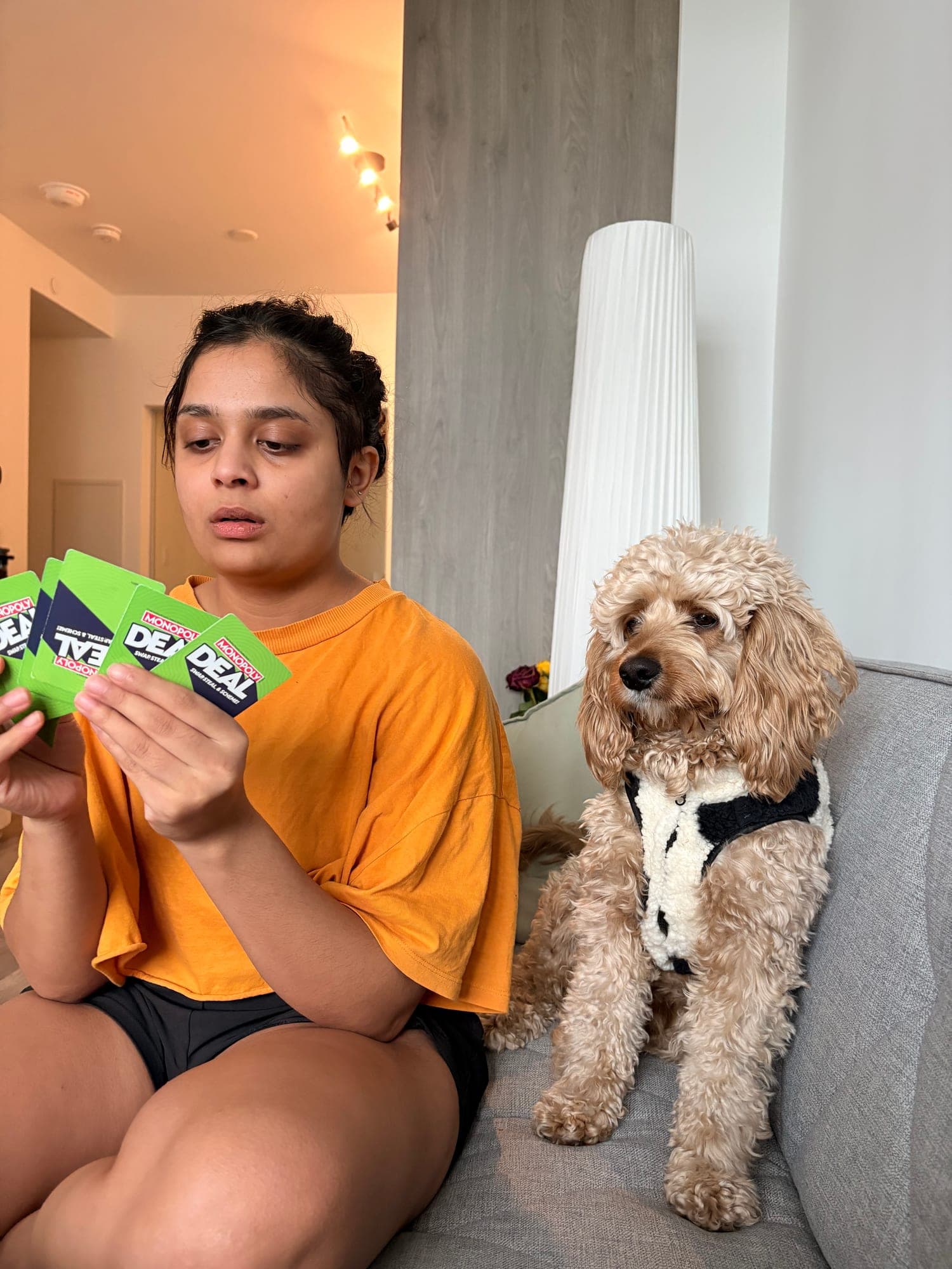 Khushi in an orange t-shirt holding up green playing cards on a grey couch, a curly apricot dog in a black-and-white harness sitting beside her.