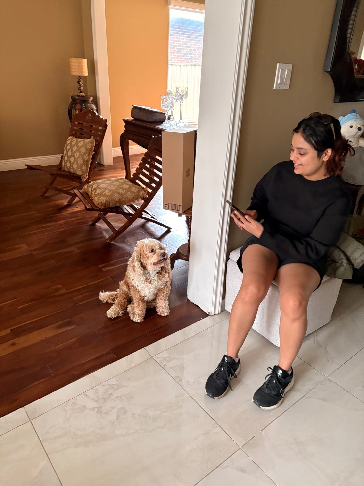 Khushi sitting by an open doorway looking at her phone, a small cream dog sitting on the wooden floor nearby.