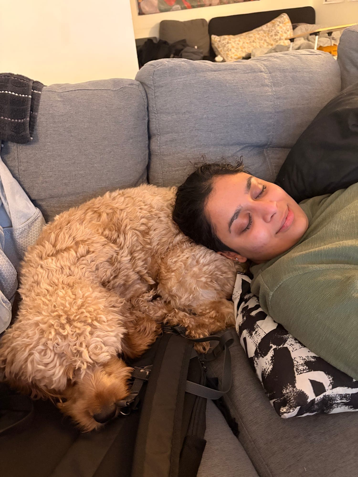 Khushi lying on a grey couch with her eyes closed, a curly apricot dog curled up asleep against her shoulder.