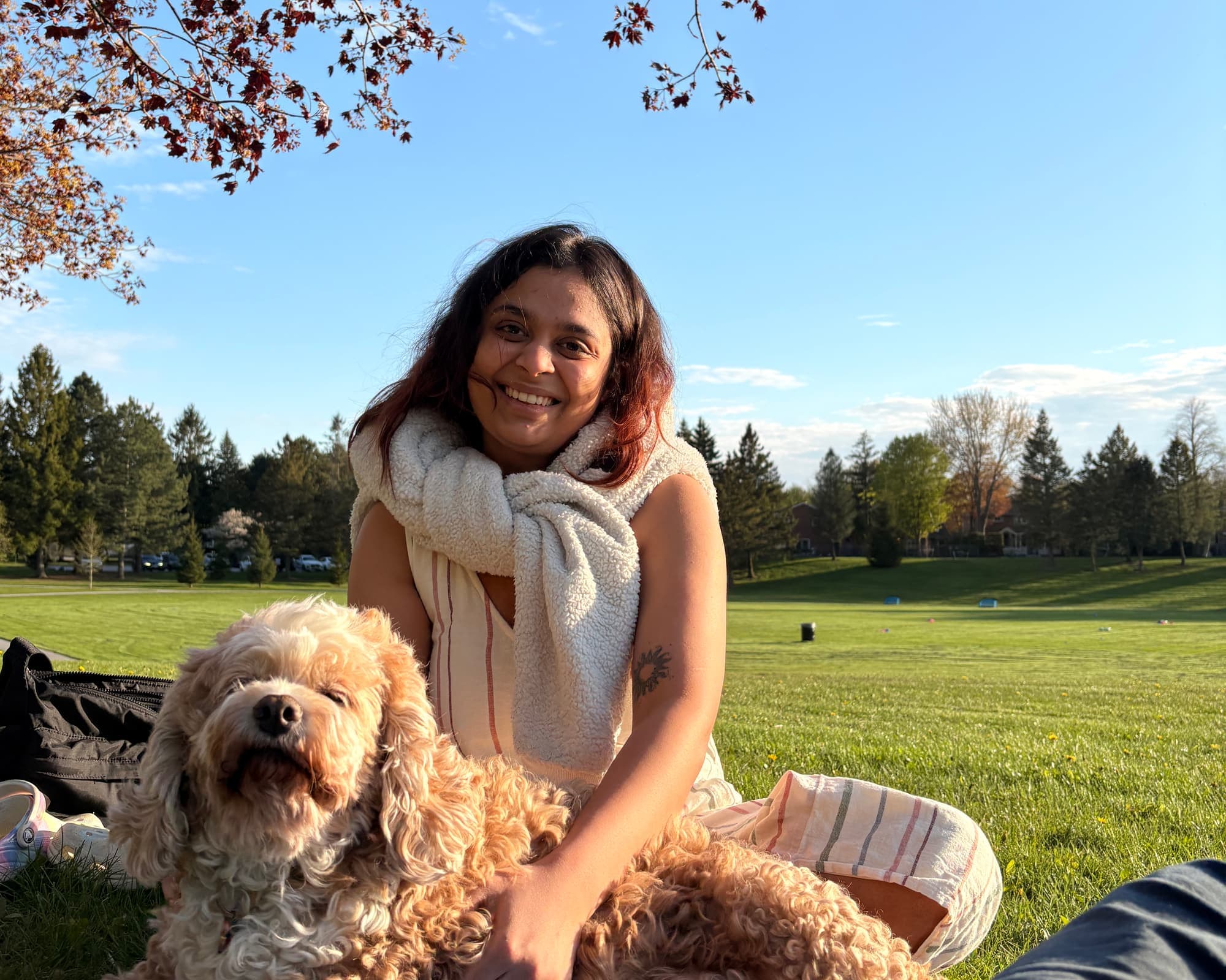 Khushi sitting on a picnic mat in a park under a blossoming tree, smiling, with a curly apricot dog stretched out in front of her and her hand on its back.