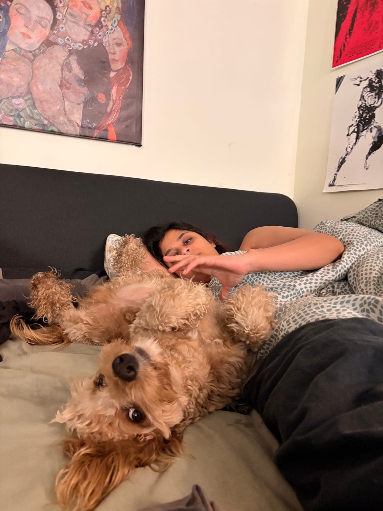 The same apricot dog lying upside down on the bed with its head toward the camera, Khushi lying behind it with a hand raised.