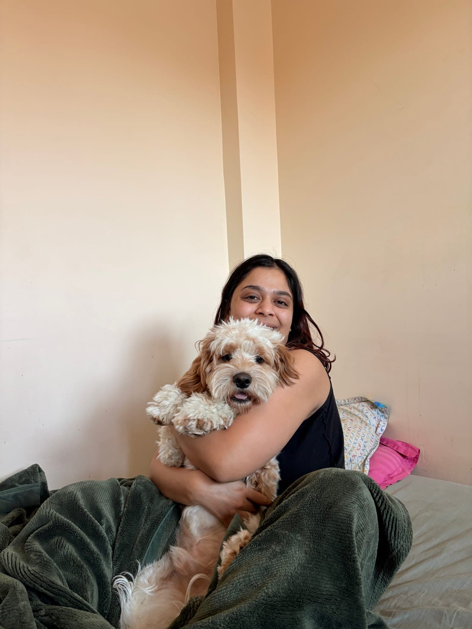 Khushi sitting on a bed, laughing and hugging a fluffy cream-and-apricot dog to her chest.
